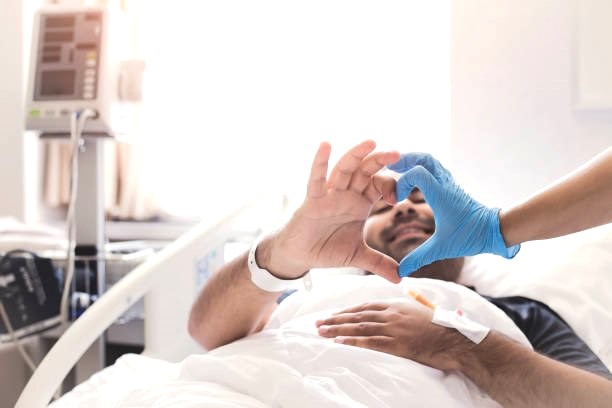 a man in a hospital bed making a heart with the hand of medical staff wearing a blue glove